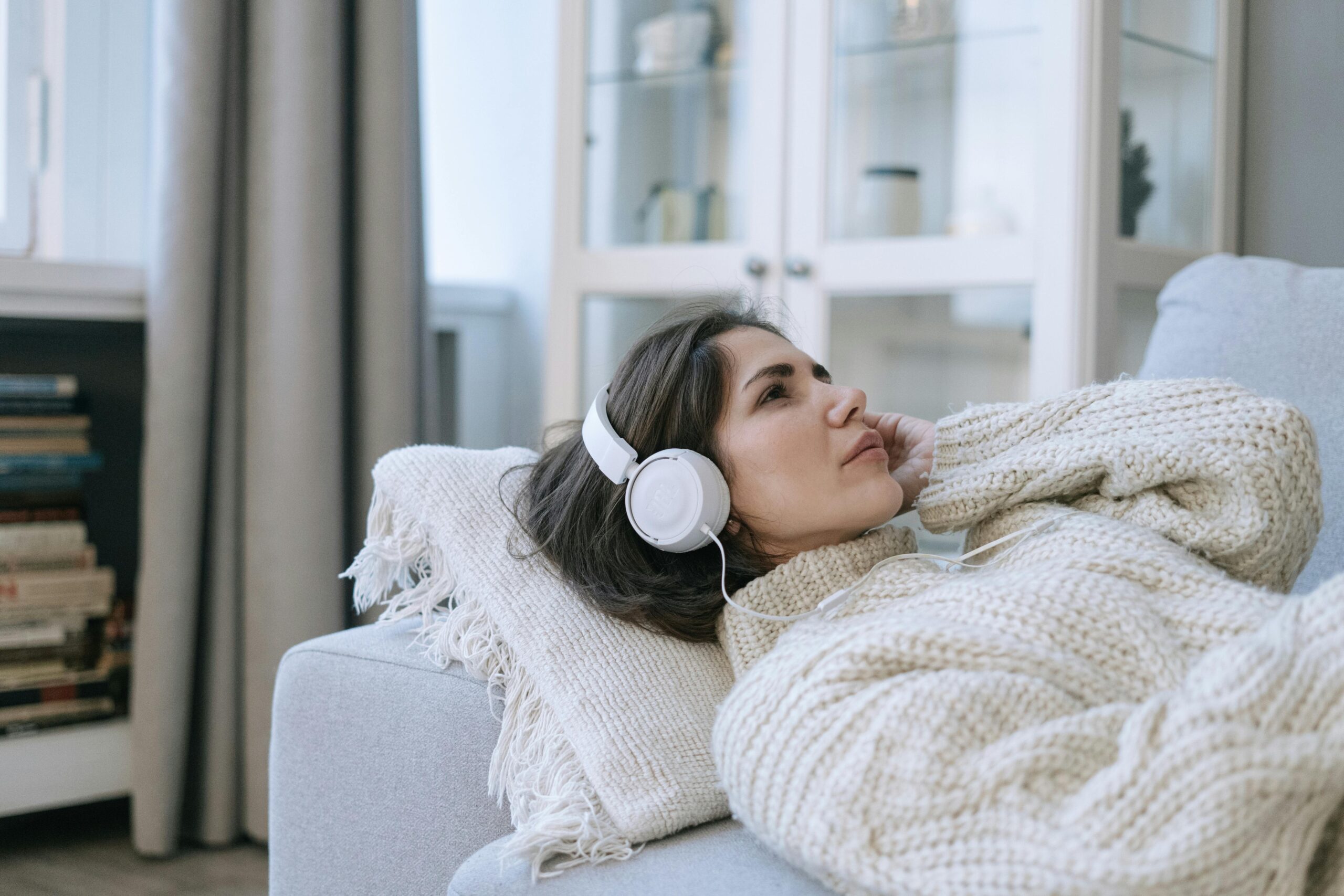 Woman in a knitted sweater relaxing on sofa, enjoying music with headphones indoors.